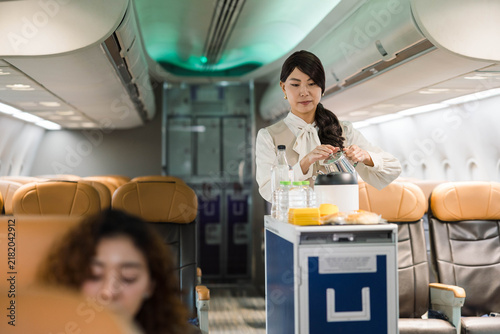 Konstfotografi Flight attendant prepares drinks on a service cart in a modern airplane cabin, with leather seats and a passenger in the foreground, illustrating professional inflight service