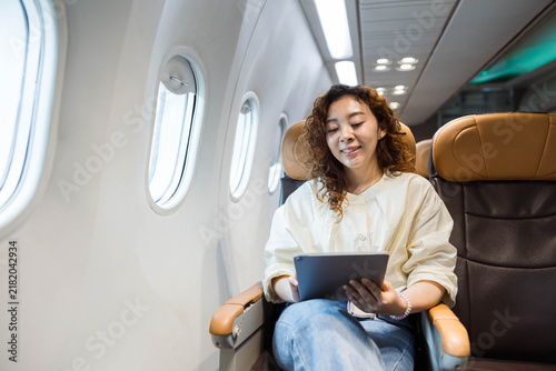 Konstfotografi Smiling passenger using a tablet in an airplane window seat, lit by cabin lights during flight, conveying leisure travel, inflight entertainment, and a modern connected lifestyle