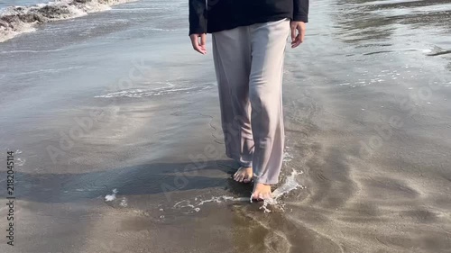 The footsteps of a woman wearing trousers walk forward among the small waves that foam on the sandy beach in the morning.