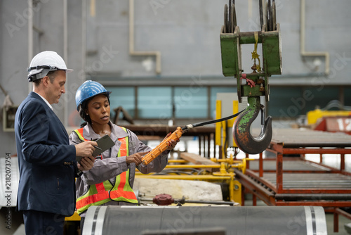 Owner boss manager assistant secretary inspection inspecting in warehouse inventory storage Female supervisor holding pendant remote controller operating overhead crane hook beside male manager