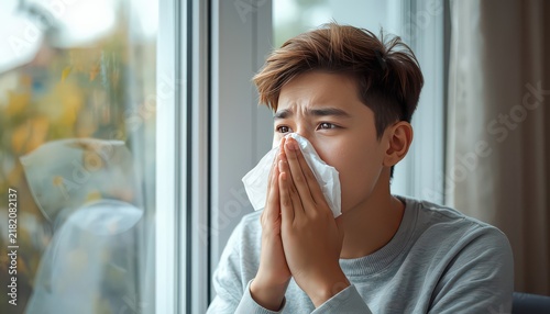 Young man blowing his nose with a tissue by a window