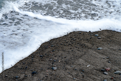 Konstfotografi Gentle white ocean wave foam washing over dark volcanic sand beach, natural coastal tide line background