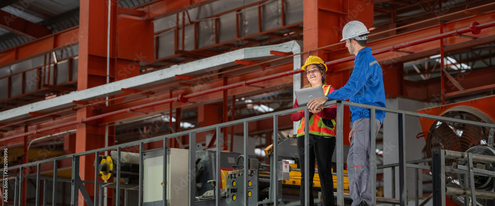 © BESTIMAGE - Engineers team discussing metal sheet machine system operation collaboration in factory Industrial male and female engineers leaning metal railing with laptop on elevated factory catwalk