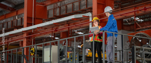 Engineers team discussing metal sheet machine system operation collaboration in factory Industrial male and female engineers leaning metal railing with laptop on elevated factory catwalk