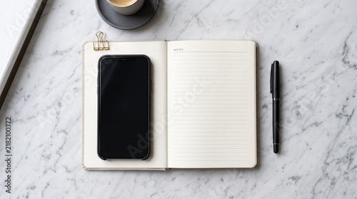 Elegant composition of black smartphone resting on open lined notebook, gold binder clip, sleek black pen, cup of espresso on grey saucer, white marble table surface