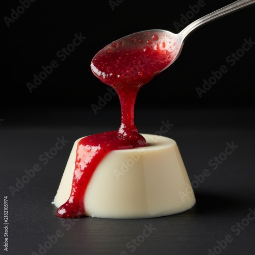 Red raspberry or strawberry sauce pouring from a spoon onto a white panna cotta dessert