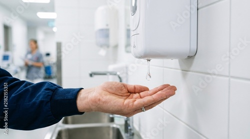 Clear drop of hand sanitizer falling from a white automatic wall dispenser into a waiting cupped hand, clean clinical healthcare setting
