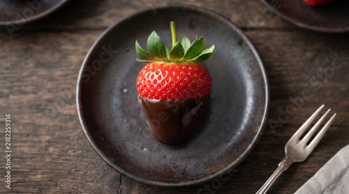 Ripe red strawberry glazed in dark chocolate on a rustic grey ceramic plate, set upon weathered wooden table with soft natural lighting.