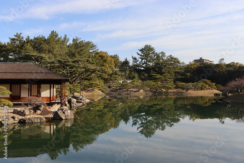 Cuadro en lienzo Traditional Japanese garden with a wooden teahouse and pine trees reflecting in a calm pond under a clear blue sky