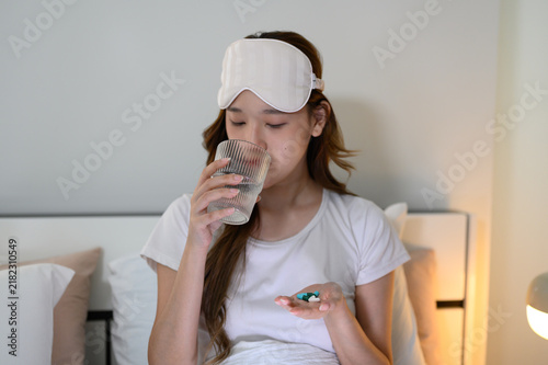 Картината върху платно Young Asian woman wearing an eye mask sitting on bed at night, holding pills in hand and drinking water