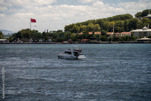 Photography Small white motor boat sails on blue sea water surface near green coastal hill with waving turkish flag