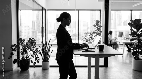 Silhouette of professional businesswoman working on laptop at standing desk in modern office with city view, focusing on productivity, remote work, corporate lifestyle