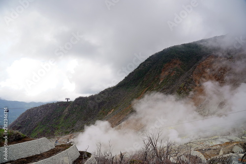 Foto 箱根大涌谷の火山活動により噴煙が上がる山肌の風景。ロープウェイが往来し、荒涼とした大地と自然のエネルギーが共存する様子。