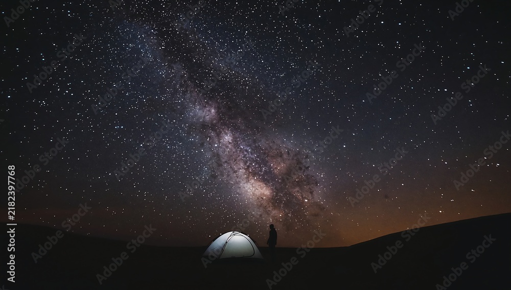 Fototapeta premium Lone camper standing by a glowing tent under a bright starry night sky and milky way