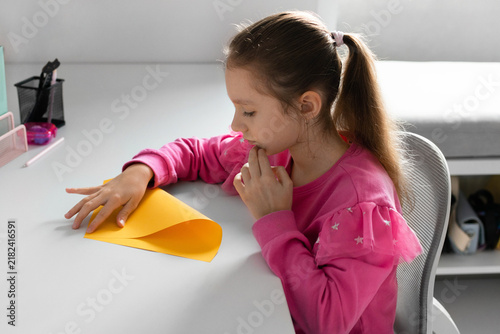 A focused young girl sits at her desk, carefully folding a yellow sheet of paper to make origami during a creative lesson at home