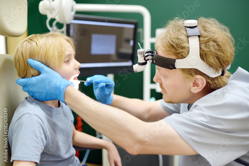 An experienced otolaryngologist examines a patient's throat using a medical tongue depressor in the consulting room. The professional ENT specialist is at their workplace.