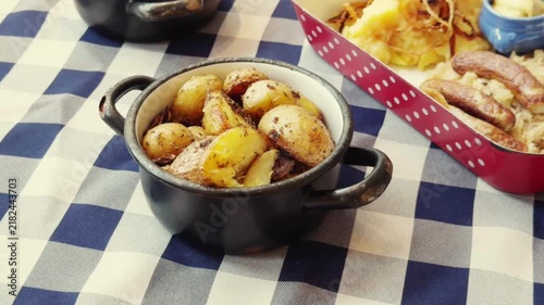 Roasted baby potatoes in black pot, traditional German sausages with sauerkraut, rustic picnic food on blue checkered tablecloth