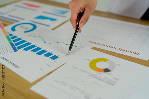 Business analyst pointing pen at growth chart on desk covered with financial reports, sales graphs and pie charts, analyzing annual data with calculator nearby