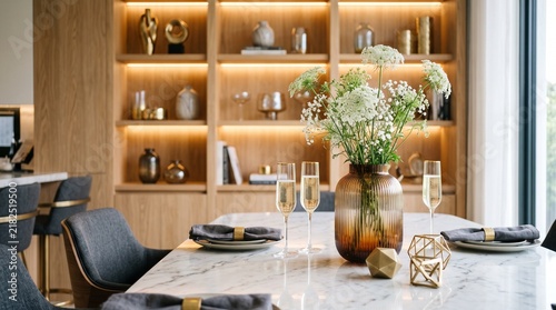 Luxury Dining Room Interior Featuring A Marble Table With Champagne Flutes And A Floral Arrangement Set In Front Of Wooden Shelves With Ornaments Creating A Warm Inviting Atmosphere For Social
