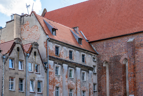 Quadro su tela Old weathered tenement facade showing damaged plaster with exposed red bricks, steep roof with red ceramic tiles, dormer windows, directly adjacent to a gothic brick church structure