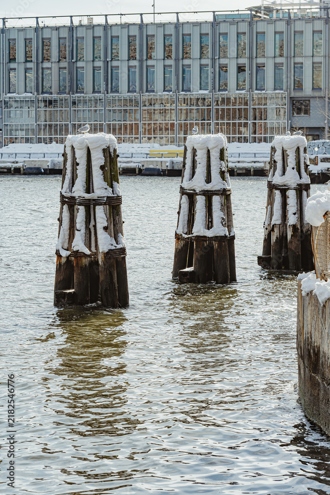 Fototapeta premium Snowy wooden piling dolphins in East River at New York. Three timber mooring posts stand in water near waterfront building.