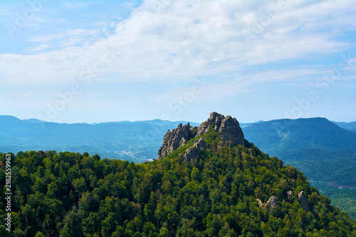 Rocky peak rising above a dense green forest. Blue mountains in a haze in the background. Clear sky with clouds.