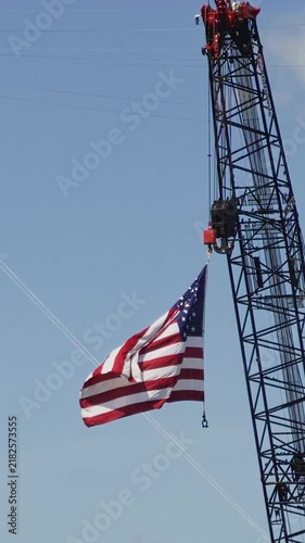 Large united states flag hanging from a tall construction crane jib and waving slowly in the wind against a clear sky. A patriotic symbol