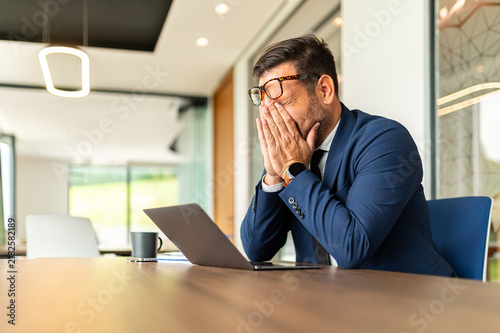 Obraz na plátně Businessman at his desk rubbing temples, eyes strained and exhausted over laptop