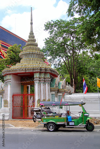 A traditional green Thai tuk-tuk auto-rickshaw parked on the street outside the entrance gate and ornate spires of Wat Pho temple in Bangkok, Thailand.