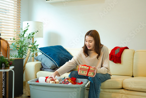 Smiling Asian woman on couch holds wrapped holiday gifts and Christmas decoration unpacked from a storage bin at home, ideal for Christmas preparation and seasonal lifestyle content