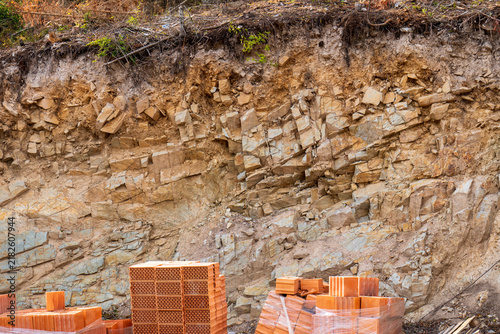 Foto Stacks of fired hollow clay bricks stored below an exposed rocky excavation wall at a construction site, showing building materials, geology and ground preparation