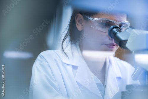 Obraz na plátně A woman lab technician in white laboratory clothing, safety glasses and blue gloves studying specimen through binocular head in a bright modern life science laboratory