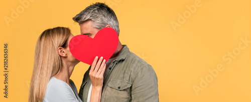 Fotografie Happy Couple, Caucasian, Adult, Showing Affection By Kissing While Holding A Red Heart In Front Of Their Face, Set Against A Bright Yellow Background