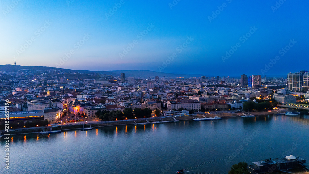 Obraz premium Panoramic skyline of Bratislava along Danube river during blue hour. Deep sky extends over illuminated embankments and city buildings.