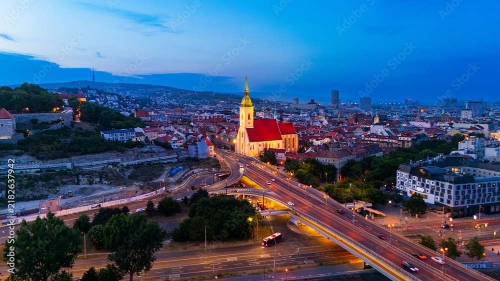 Fototapeta premium St Martin Cathedral and illuminated highway traffic at night in Bratislava. Cathedral tower glows gold near busy highway overpass with red car light trails.