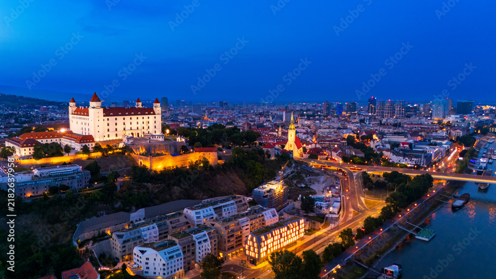 Obraz premium Bratislava city view with castle at dusk showing river and buildings. Bratislava shows its skyline at dusk with lights on buildings and the river reflecting them.