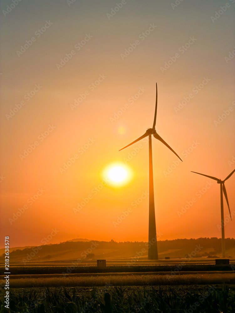 Obraz premium Sunset over wind turbines in a rural landscape. The sun sets behind wind turbines in a rural area. The sky shows orange and yellow colors.