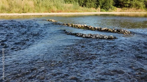Clear, clean mountain river is flowing in the shallows, low-head dam across the river, riverbank with grass and trees, natural background