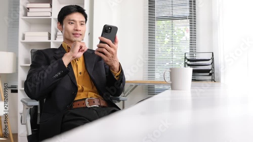 Smiling Asian businessman using a smartphone in a modern office, sitting at his desk and scrolling through his phone. He is relaxing during a work break in office.