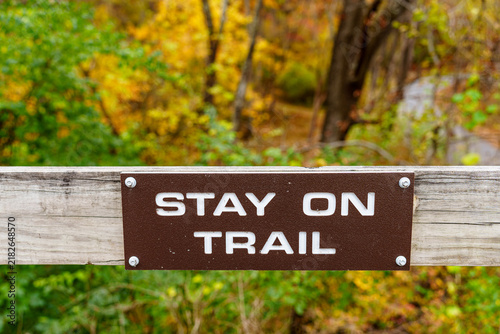 Sign instructing to stay on trail in an outdoor setting surrounded by trees and autumn leaves