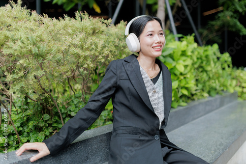 Smiling businesswoman wearing headphones in a modern green office courtyard, showing professional lifestyle, relaxation, technology, wellbeing, confidence, and healthy work-life balance.