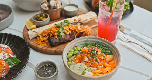 Fotografie Fresh salmon poke bowl with edamame and vegetables served next to a grilled meat platter and pink cocktail