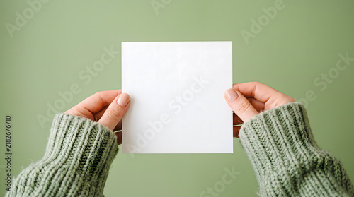 Cozy autumn Thanksgiving mockup with hands wearing green knitted sweater holding blank square white paper card on matching green background empty space for text
