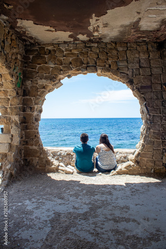 Canvastavla Couple sitting viewing Mediterranean Sea through old stone arch