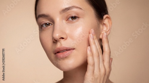 A young woman with glowing skin touching her cheek gently against a neutral beige background with natural makeup touching