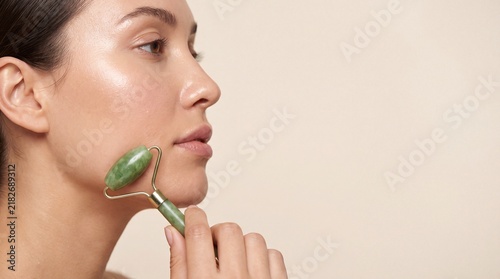 Young woman rolling green stone massager tool along cheek for facial skincare routine with roller tool her