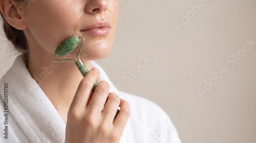 Woman using a jade facial roller on her jawline while wearing a white bathrobe indoors with bathrobe uses green