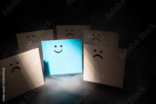 Obraz na plátne Glowing blue smiling sticky note surrounded by sad white notes in dramatic smoke on dark background
