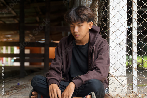 Sad Asian teenage boy sitting on ground against a chain-link fence, depicting youth depression, isolation, and stress.