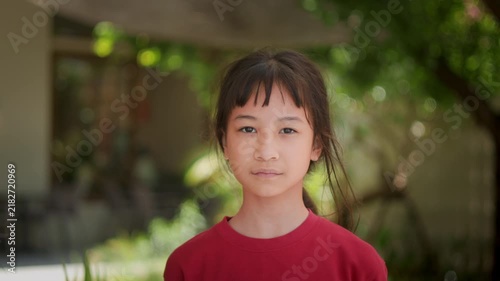 Portrait of adorable Asian kid looking directly at the camera with a gentle smiling face in a red shirt standing outdoors with beautiful green bokeh background. Childhood happiness lifestyle concept.